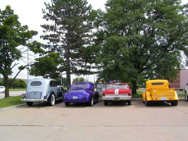Row of restored customer cars — a blue classic sedan, purple street rod, red Fairlane and yellow hot rod pickup — parked at the shop